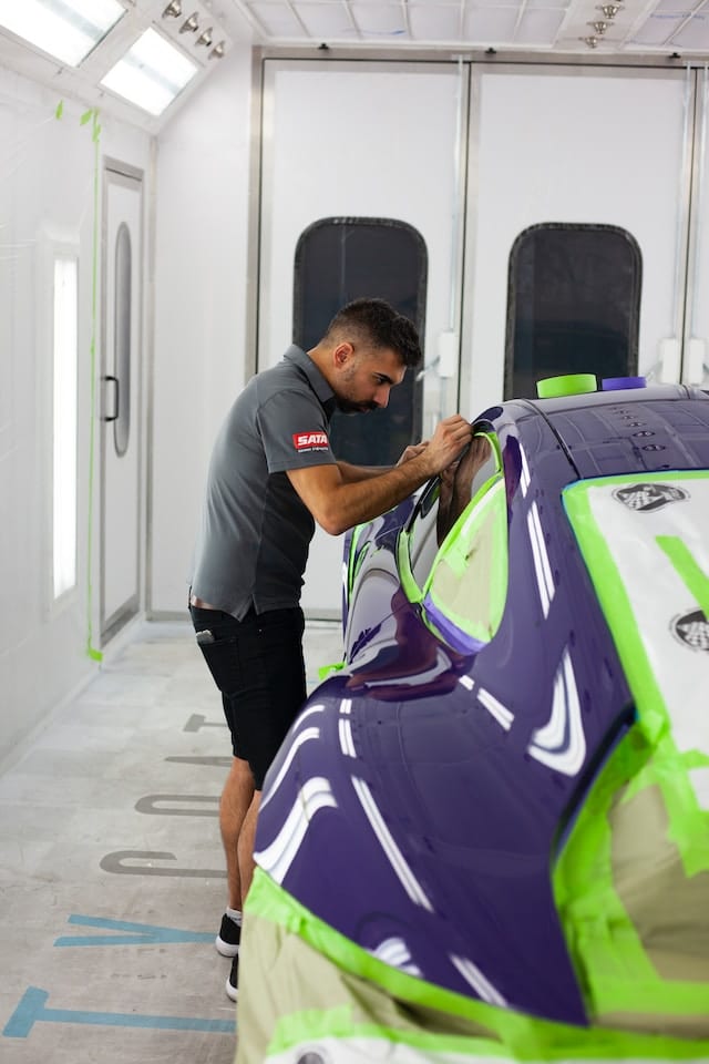 Worker meticulously applying green masking tape to the edges of a sports car's windows, preparing the vehicle for a paint job.