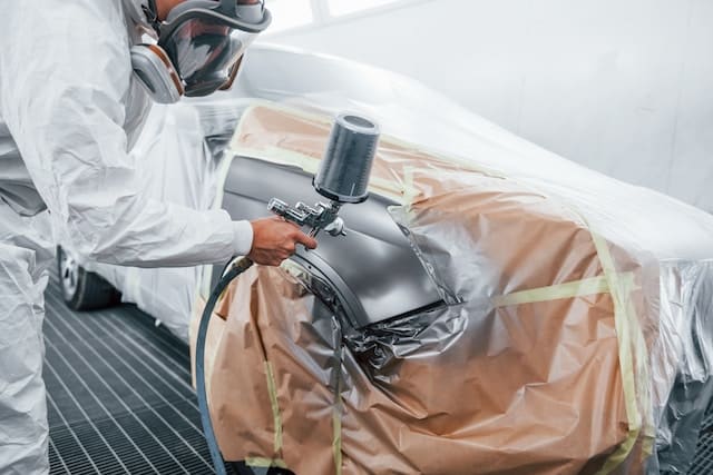 Worker in a protective hazmat suit skillfully painting the right front section of a car with a gun sprayer, applying a sleek grey metallic color.