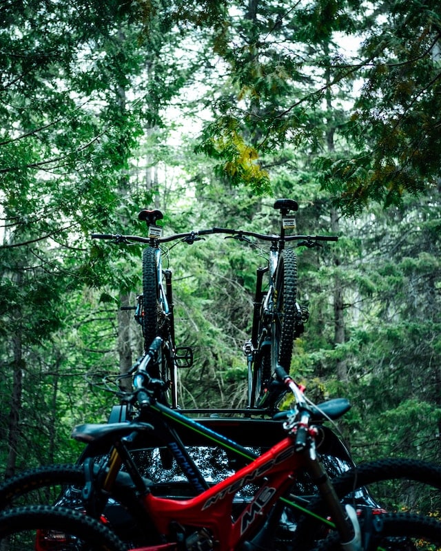 Close-up of a car equipped with two bikes mounted on the roof and another two bikes secured at the back, set against a serene forest backdrop.