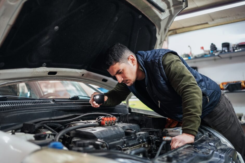 An image of a mechanic attentively inspecting a car for jerking issues, signifying the need for professional intervention. The mechanic is shown peering under the hood, with a look of concentration, holding tools that suggest a detailed examination is underway. This photo underscores the complexity of car jerking problems and the expertise required to diagnose and fix them. The background of the garage, filled with automotive equipment, reinforces the professional setting necessary for such technical evaluations.