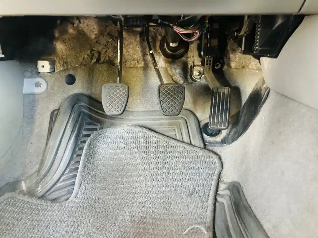 Photograph of three pedals from a manual transmission car, focusing on the clutch pedal on the left. The clutch pedal shows signs of wear, indicative of age and use, potentially causing a squeaking noise when depressed.