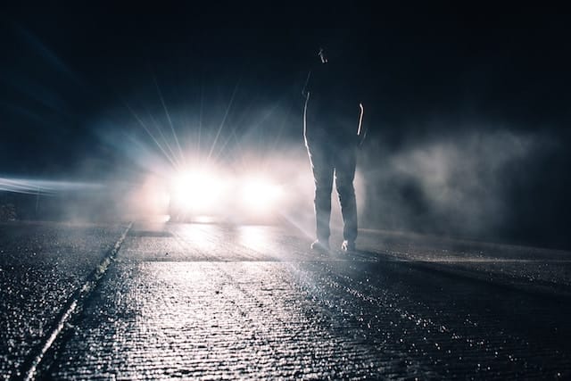 A car with its headlights on facing a man standing in the middle of a country road at night, the surrounding darkness enveloped by the vehicle's bright beams.