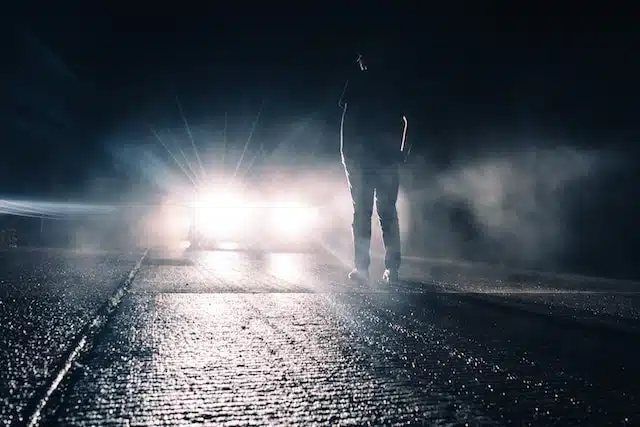 A car with its headlights on facing a man standing in the middle of a country road at night, the surrounding darkness enveloped by the vehicle's bright beams.