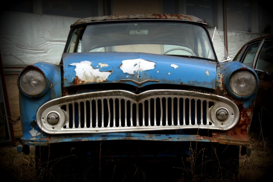 A close-up image of a dilapidated car showcasing signs of significant wear and tear. The car's paint is faded and peeling, with visible rust and dents covering its body. Broken windows and a partially detached bumper further emphasize the car's abandoned and decayed state.