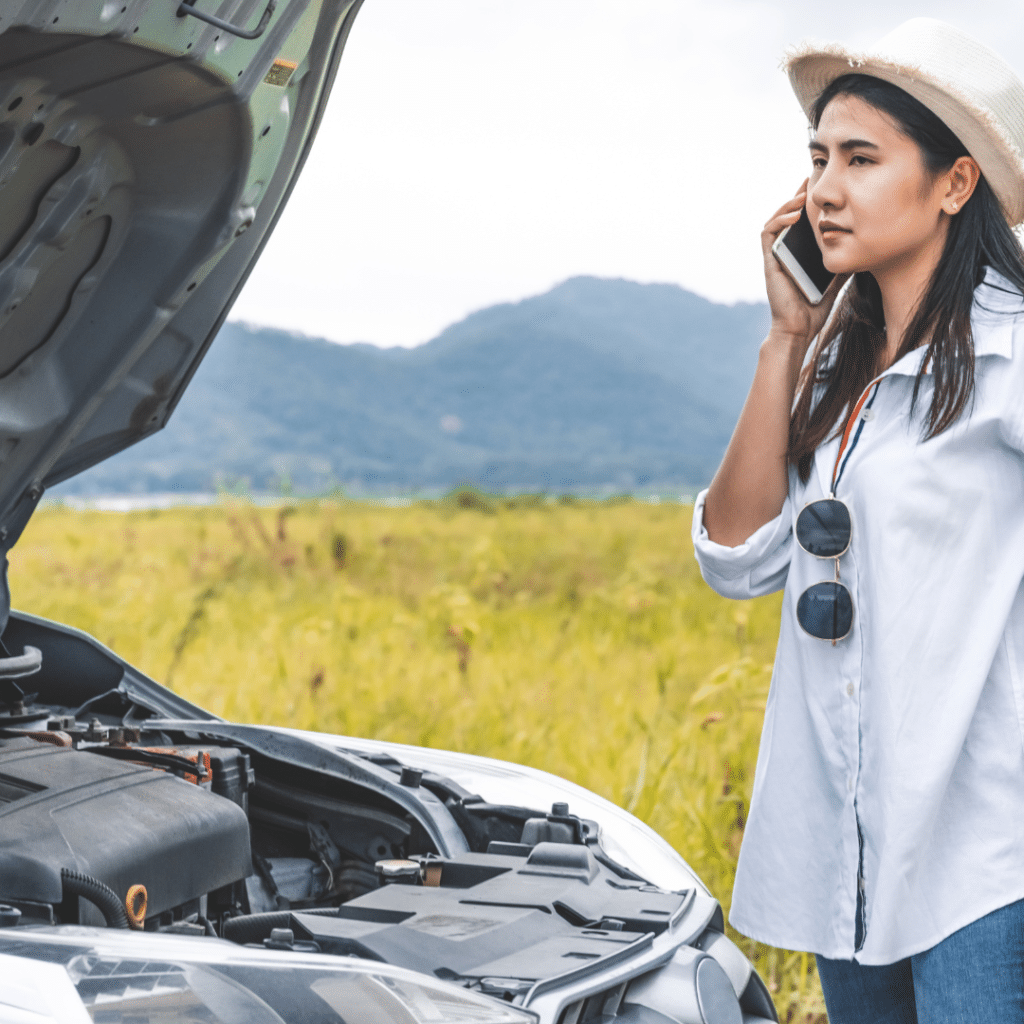 A woman stands by her stranded car on the roadside, speaking on her mobile phone with a look of concern, seeking assistance